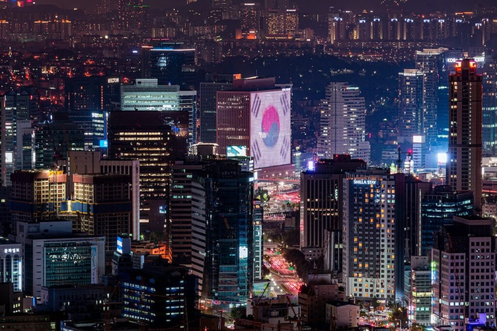 Aerial view of downtown Seoul lit up at night, with mid- and high-rise buildings, traffic below, and an image of the South Korean flag projected onto one of the office buildings.